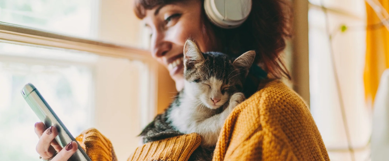 a woman wearing headphones with a kitten on her shoulder looking at her mobile phone