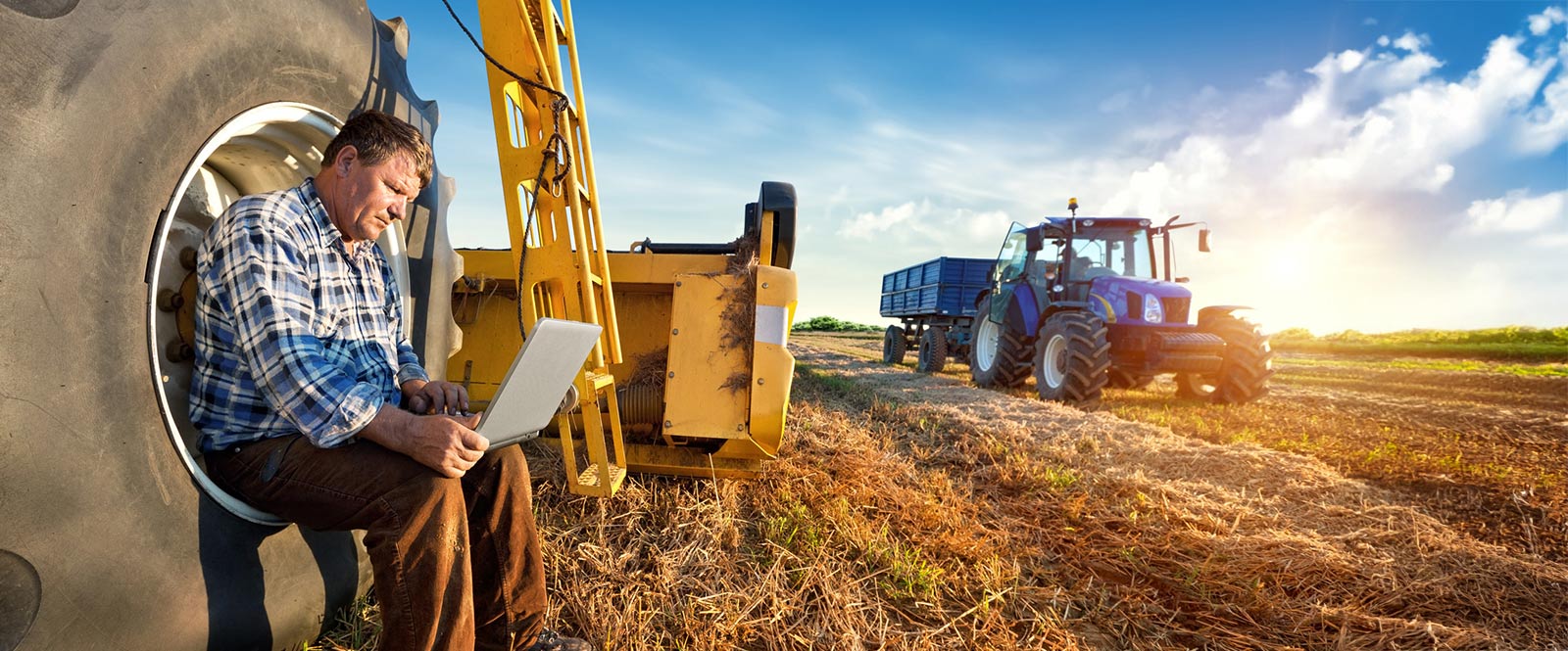 Farmer sitting on tractor wheel using a laptop.